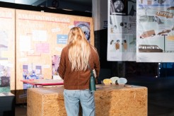 Eine Frau mit langen, blonden Haaren und brauner Jacke steht vor einer Ausstellungstafel mit dem Titel 'Stadtentwicklung'. Auf der Tafel sind verschiedene Schaubilder, Notizen und Informationen zur Stadtplanung angebracht. Im Hintergrund sind weitere Poster und Schautafeln zu sehen.