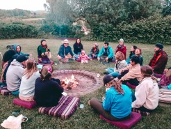 Eine Gruppe von Menschen sitzt auf Kissen um ein Lagerfeuer auf einer Wiese. Sie sind in warme Kleidung gehüllt und scheinen in ein Gespräch vertieft zu sein. Im Hintergrund sind Bäume und Sträucher zu sehen, die die gemütliche Szenerie umrahmen.