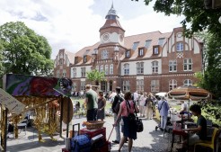 Das historische Backsteingebäude vom Coworkhaus im Anscharpark Kiel liegt im Hintergrund. Davor findet ein Flohmarkt mit vielen umherziehenden Menschen statt.