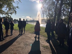 Projektmitarbeitende von Liveability stehen in einem Park in Pori, Finnland. Im Hintergrund sprudelt eine Wasserfontäne. Die Sonne scheint und die Körper Menschen werfen lange Schatten auf einen Weg.