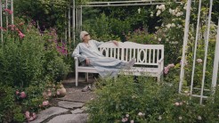 Filmstill aus Ihr Jahrhundert. Eine ältere Frau mit langem Kleid und Sonnebrille sitzt auf einer weißen Bank. Die Bank ist von einem malerischen Garten umgeben.