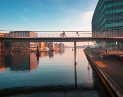 Eine moderne Fußgänger- und Radfahrerbrücke erstreckt sich über einen ruhigen Kanal in einer städtischen Umgebung. Auf der Brücke ist eine Person zu sehen, die mit einem Fahrrad fährt. Die umliegenden Gebäude reflektieren das Licht des Sonnenuntergangs und spiegeln sich im Wasser des Kanals. Das Bild vermittelt eine ruhige und idyllische Atmosphäre.