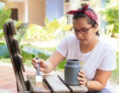 Eine Perseon sitzt vor einem Stuhl und bemalt diesen neu. Es handelt sich hierbei um Upcycling.