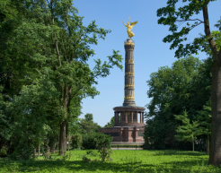 Die Berliner Siegessäule steht majestätisch inmitten des grünen Großen Tiergartens, umgeben von üppigen Bäumen und einer gepflegten Rasenfläche. Die goldene Statue der Viktoria thront auf der Spitze der Säule und leuchtet im Sonnenlicht.