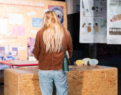 Person mit blonden langen Haaren steht mit dem Rücken zur Kamera vor einem Pult. Im Hintergrund befindet sich eine Pinnwand mit zahreichen Flyern, Postern und weiteren Ausstellungsinformationen.