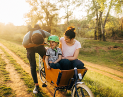 Eine Erwachsene Person sitzt mit einem Kind in einem Lastenrad. Gefahren wird es von einer weiteren erwachsenen Person. Im Hintergrund ist ein Feldweg und der Sonnenuntergang zu sehen.