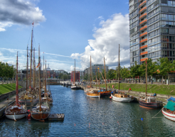 Der Kieler Hafen. Zu sehen sind mehrere kleine Segenschiffe in einer kleinen Hafenbucht. Im Hintergrund sind die Stadt und ein hohes, gläsernes Gebäude zu sehen.