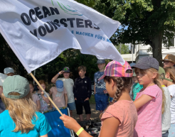 Eine Gruppe von Kindern steht im Kreis zusammen. Ein Kind hält eine Fahne mit der Auschrift "Ocean Youngsters" in der Hand. Die Kinder stehen draußen. Im Hintergrund sind Bäume zu erkennen.