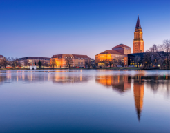 Ein beleuchtetes Stadtpanorama von Kiel bei Abenddämmerung mit Blick auf ein ruhiges Gewässer. Die beleuchteten Gebäude und der hohe, markante Rathhausturm spiegeln sich klar im Wasser wider. Der Himmel zeigt ein sanftes Blau, das langsam in die Dunkelheit übergeht. Die Szene strahlt Ruhe und Eleganz aus.