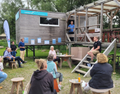 Vor einem Holz-Container sitzen einige Menschen auf Hockern und Stühlen. Sie hören einer Person zu. Diese sitz auf der Treppe, die zu dem höher gestellten Container führt. 