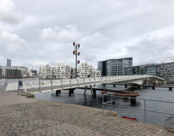 Eine moderne Fußgängerbrücke über einen Kanal. Die Brücke hat ein leicht geschwungenes Design und ist mit Metallgeländern gesichert. Im Hintergrund sind moderne Wohn- und Bürogebäude mit großen Fenstern und geometrischen Fassaden zu sehen. Der Himmel ist bewölkt, und das Wasser unter der Brücke spiegelt die Umgebung. Am Ufer ist ein gepflasterter Weg mit einem Geländer erkennbar.