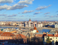 Blick auf das ungarische Parlament in Budapest, das an der Donau liegt. Im Vordergrund sind rote Ziegeldächer und historische Gebäude zu sehen, während der Himmel blau mit einigen weißen Wolken ist.