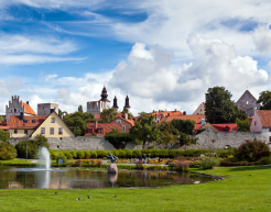 Eine malerische Stadtansicht mit historischen Gebäuden und Kirchen, die sich hinter einer Steinmauer erheben. Im Vordergrund befindet sich ein gepflegter Park mit einem Teich, in dem eine kleine Fontäne sprudelt. Der Himmel ist blau mit weißen Wolken, was dem Bild eine friedliche und idyllische Atmosphäre verleiht.