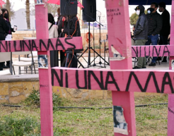 Die Aufnahme zeigt mehrere pinkfarbene Holzkreuze mit der Aufschrift „Ni una más“, was auf Spanisch „Nicht eine mehr“ bedeutet. Im Hintergrund versammeln sich Menschen, einige tragen Masken, während eine Veranstaltung mit Lautsprechern und Mikrofonen stattfindet. Das Bild verweist auf Proteste gegen geschlechtsspezifische Gewalt und Femizide, insbesondere in Lateinamerika.