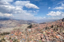 La Paz in Bolivien von oben. Unzählige Häuser stehen an einem Hang. Am Horizont sind Berge und blauer Himmel mit weißen Wolken.