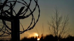 Im Vordergrund sieht man Teile eines Stacheldrahtzauns, darunter ein Zaun. Im Hintergrund ist der Sonnenuntergang verschwommen hinter einem Baum.